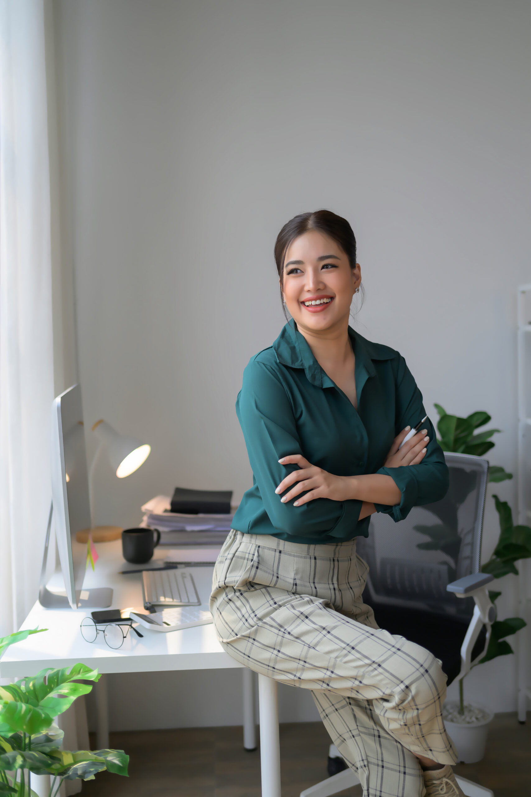 A woman is sitting at a desk with a computer monitor and a keyboard in front of her. She is wearing a green shirt and plaid pants